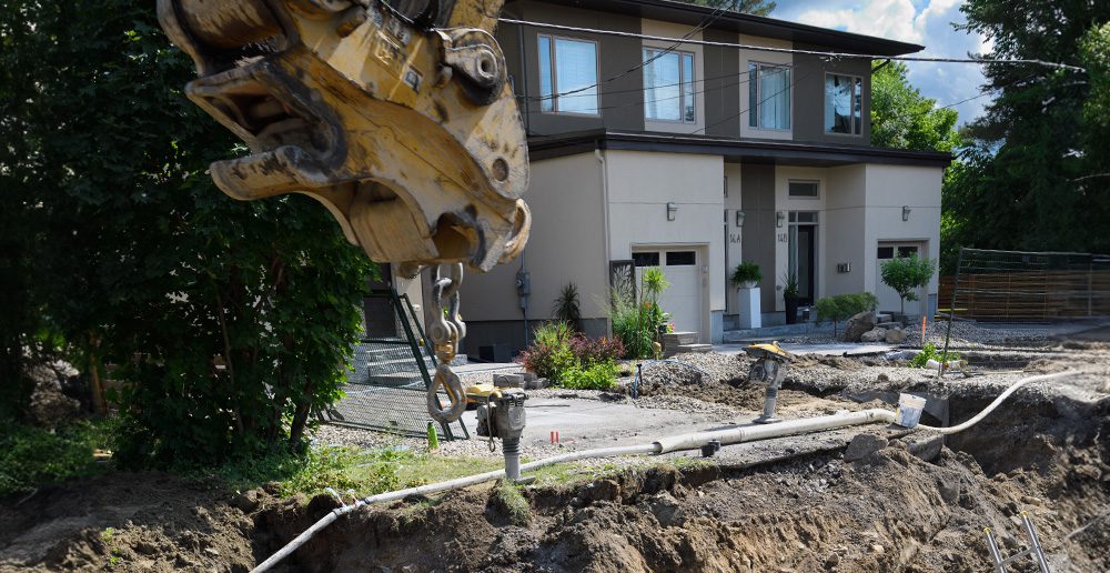 Excavator digging a trench for a sewage line in front of a house.